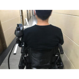 轮椅王在米色墙壁的走廊里，一名身穿黑色衬衫的人坐在配备轮椅顶部控制配件（适合型号：豪华500、豪华500 加强版及豪华1200）的电动轮椅上，面朝远处。