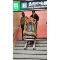輪椅王 在香港中環地鐵站出口(標示為「B」)處,兩人正在協助一名坐在電動輪椅上的人走下樓梯。輪椅被固定並在樓梯升降機上被引導。