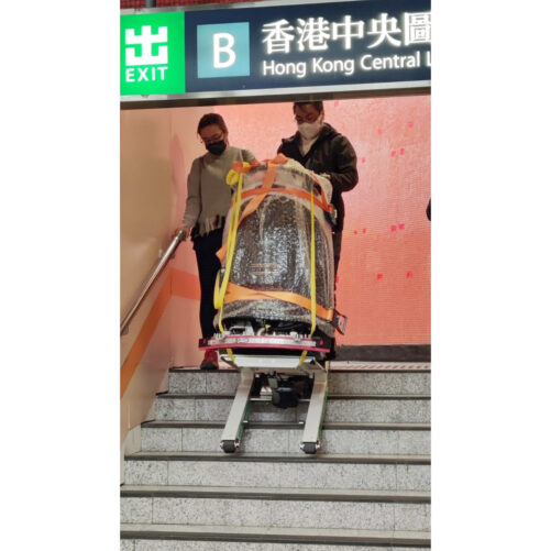 輪椅王 在香港中環地鐵站出口（標示為「B」）處，兩人正在協助一名坐在電動輪椅上的人走下樓梯。輪椅被固定並在樓梯升降機上被引導。