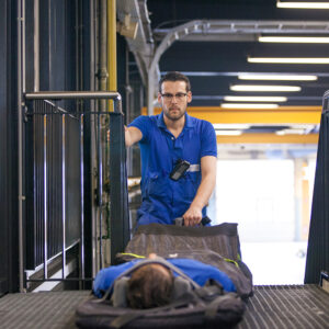Roi en fauteuil roulant Un homme en salopette bleue pousse une civière avec une autre personne allongée dessus dans un couloir de style industriel.