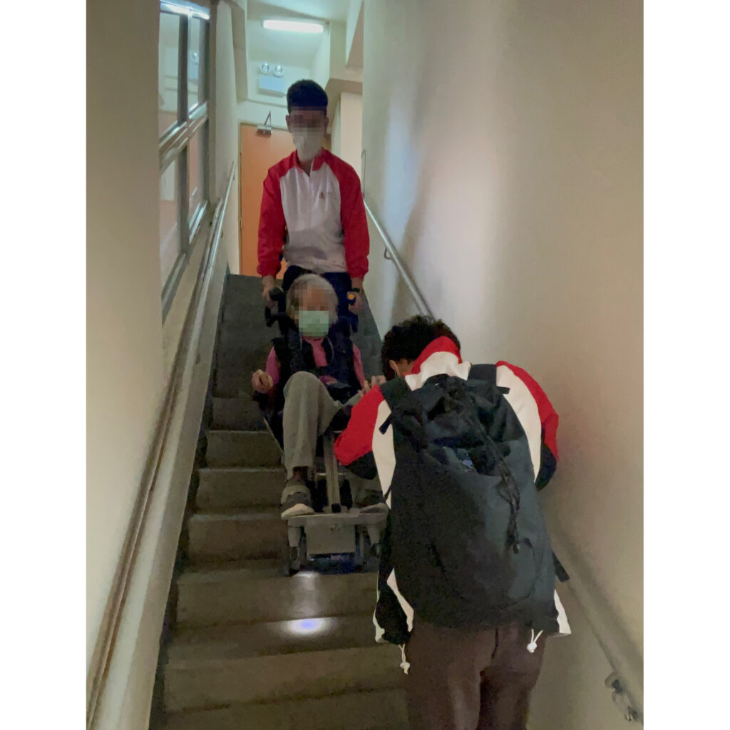 3 人が Wheelchair King 階段機械を使用して、車椅子に乗った人を建物内の階段を慎重に誘導しています。前方に1人、後方に2人が介助しており、いずれもマスクを着用していた。