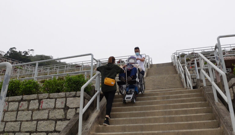El rey de las sillas de ruedas Una mujer ayuda a un hombre a empujar a otro en silla de ruedas por una larga escalera con barandillas de metal en un día nublado. Utilizaron el Wheelchair King Stairmaster para caminar con cuidado y garantizar un paso seguro.