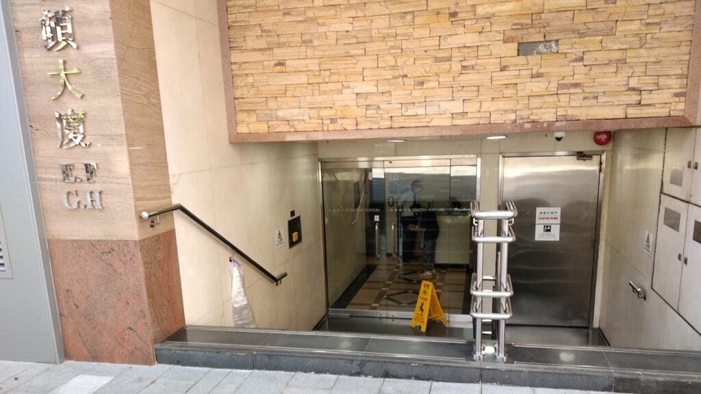 Wheelchair King の建物の入り口には、石造りのファサード、ガラスのドア、ステンレス鋼の手すりがあります。ドアの近くにある「車椅子王デモンストレーション階段マスター」の看板など、中国語と英語の看板がはっきりと見えます。床濡れ注意の標識もあります。