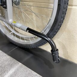 輪椅王 自行車後輪特寫，支架伸出，靠在米色牆壁的瓷磚地板上，讓人想起台灣品牌KARMA KM-2580X(B)多功能超高承重手推輪椅的堅固設計。