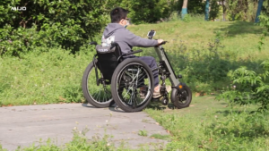 轮椅王一个坐在轮椅上的人正在户外草地上的小路上行走。他们穿着灰色连帽衫，戴着眼镜，使用MT02 手推变电动轮椅拖头，展示了旅途中的轻松和控制。
