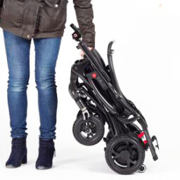 輪椅王 一個穿著牛仔褲和靴子的人站在白色背景上折疊的黑色碳纖超輕電動輪椅旁。