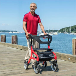 [Rey en silla de ruedas] Un hombre con una camiseta polo roja usa su andador plegable Drive Medical (rojo) para navegar por el muelle de madera, con botes y agua con gas creando un fondo pintoresco.