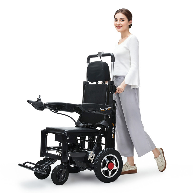 [車椅子の王様] 白いトップスとグレーのパンツを着た女性が、黒いデュアルクライマー [2-in-1 Wheelchair Stairmaster] 電動車椅子の隣に立っていました。車椅子にはジョイスティック コントロール、フットレスト、太い車輪が付いています。彼女は期待して微笑んだ。