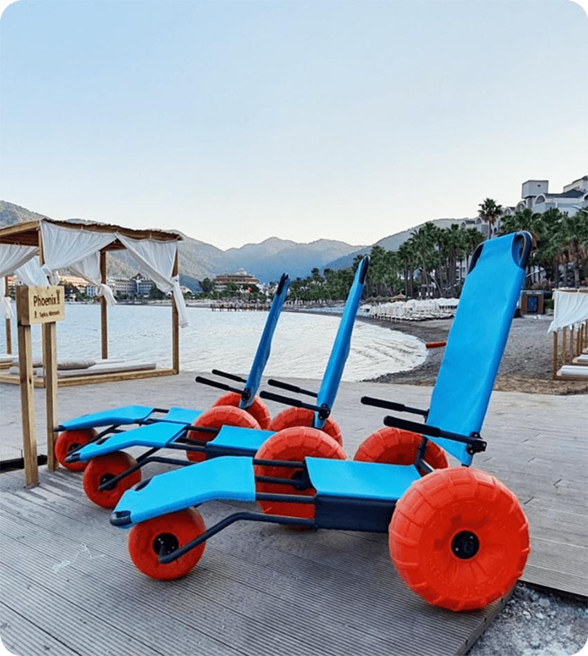 【轮椅王】 两把蓝色的Beach Star 沙滩轮椅配有橙色的大轮子,放置在海滩附近的木板路上,以山脉为背景。它们在土耳其制造,由轮椅王独家代理独家经销,承诺无与伦比的沿海机动性。