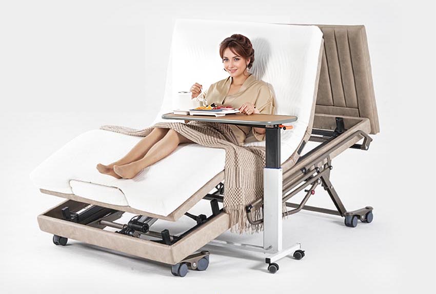 [Rey en silla de ruedas] Una mujer sentada en una cama de hospital ajustable equipada con tecnología de cama de enfermería multifuncional de cama inteligente de la marca británica Manhattan. Mientras disfruta de su comida en la bandeja, admira la comodidad del colchón TPEE+ y la comodidad del elevador de cama equipado con el control WCN931-Y (control remoto inalámbrico).