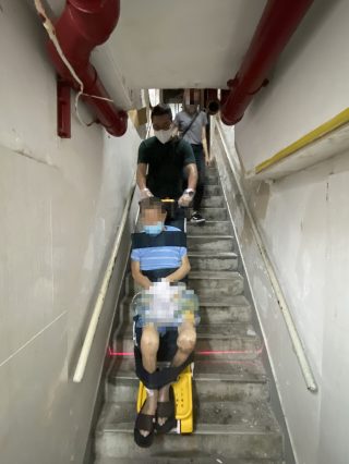 Rey en silla de ruedas Un hombre con una camisa a rayas azules es ayudado por otro hombre con una camisa verde mientras desciende de un salvaescaleras. Hay otra persona en las escaleras de entrada. Las paredes son blancas y hay tuberías rojas en lo alto, lo que muestra la dedicación de los voluntarios del Rey en Silla de Ruedas para subir y bajar escaleras a los ancianos.