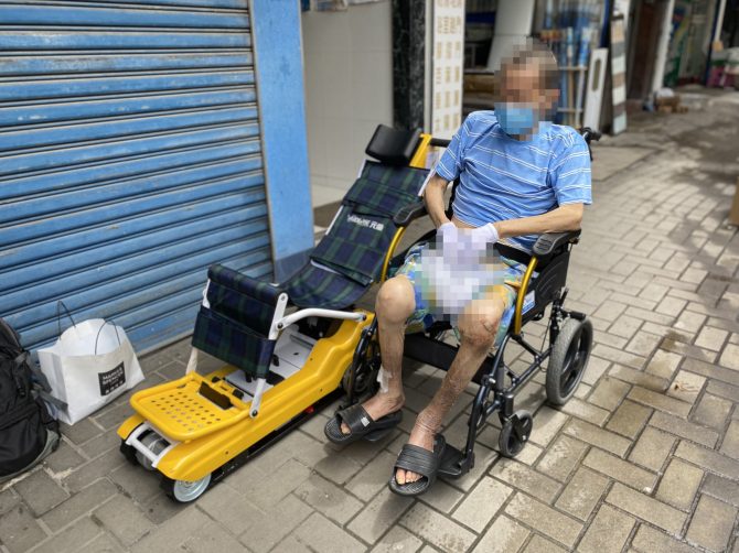 Rey en Silla de Ruedas Un hombre en silla de ruedas está sentado en la acera con un dispositivo de movilidad amarillo a su lado, que recuerda a los voluntarios del Rey en Silla de Ruedas que suben y bajan escaleras a los ancianos. Llevaba una camisa a rayas azules y sandalias, y su rostro estaba borroso.